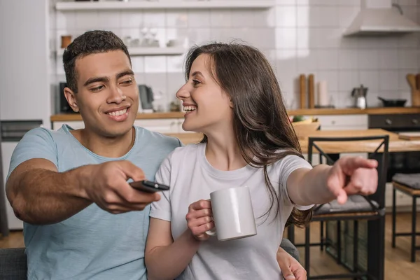 Selective focus of happy woman holding cup, pointing with finger and looking at mixed race boyfriend with remote controller — Stock Photo
