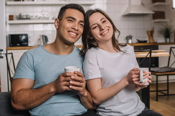 Alegre pareja interracial sosteniendo tazas de café - foto de stock