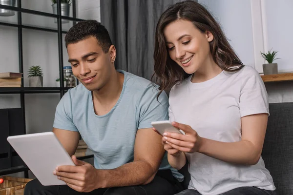 Alegre interracial pareja usando gadgets en sala de estar - foto de stock