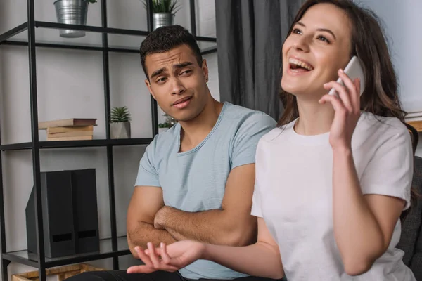 Selective focus of displeased mixed race man with crossed arms looking at girlfriend talking on smartphone — Stock Photo