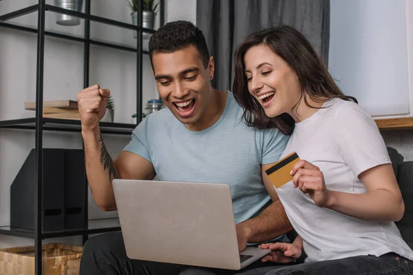 Menina feliz segurando cartão de crédito perto de namorado de raça mista olhando para laptop — Fotografia de Stock