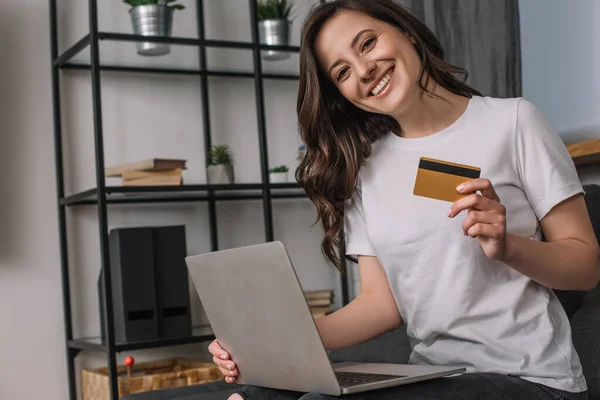 Mujer joven y feliz sosteniendo la tarjeta de crédito cerca del ordenador portátil - foto de stock