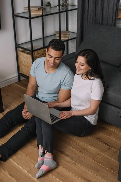 Веселая межрасовая пара, сидящая на полу рядом с диваном и использующая ноутбук — стоковое фото
