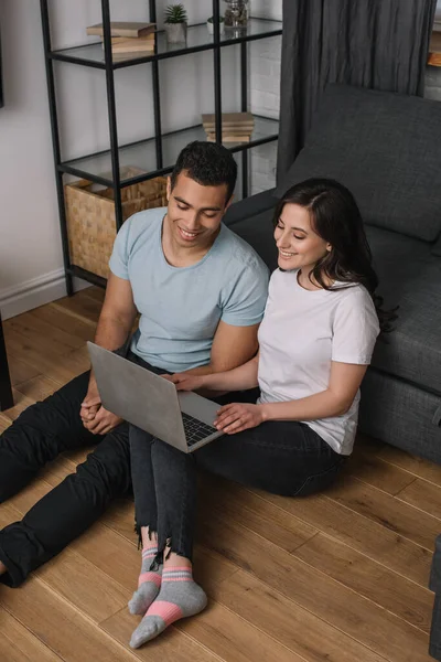 Alegre interracial casal sentado no chão perto do sofá e usando laptop — Fotografia de Stock