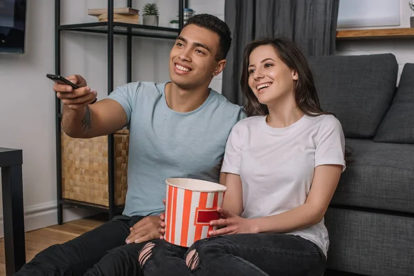 Happy mixed race man holding remote controller near cheerful girl and popcorn bucket — Stock Photo