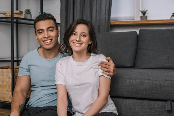 Feliz mixto raza hombre abrazando atractivo y sonriente chica en casa - foto de stock