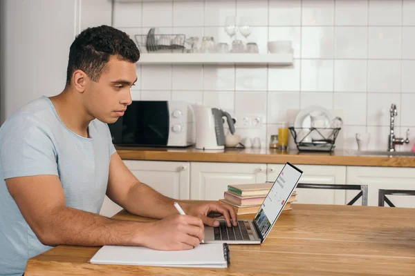 KYIV, UKRAINE - MAY 14, 2020: handsome mixed race man holding pen near notebook and using laptop with airbnb website — Stock Photo