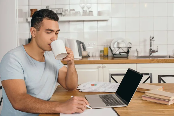 Apuesto hombre de raza mixta celebración de pluma y beber café cerca de la computadora portátil con pantalla en blanco - foto de stock