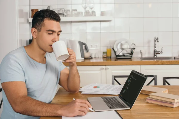 QUIIV, UCRÂNIA - MAIO 14, 2020: bonito homem de raça mista segurando caneta e bebendo café perto de laptop com javascript — Fotografia de Stock