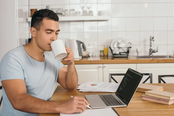 KYIV, UCRANIA - 14 de mayo de 2020: apuesto hombre de raza mixta sosteniendo la pluma y bebiendo café cerca de la computadora portátil con javascript - foto de stock