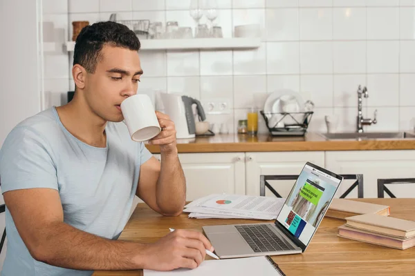 KYIV, UCRANIA - 14 de mayo de 2020: apuesto hombre de raza mixta sosteniendo la pluma y beber café cerca de la computadora portátil con sitio web bbc - foto de stock