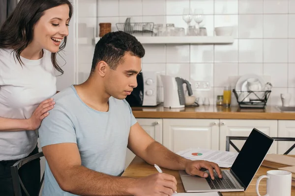 Cheerful woman standing near mixed race boyfriend holding pen near laptop with blank screen — Stock Photo