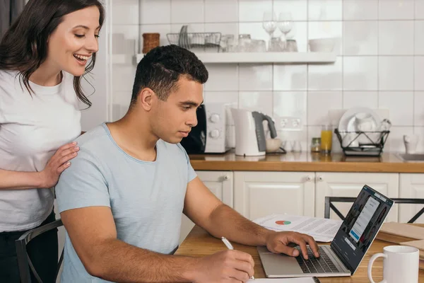 KYIV, UCRAINA - 14 MAGGIO 2020: donna allegra in piedi vicino al fidanzato di razza mista che tiene la penna vicino al computer portatile con il sito web linkedin — Foto stock