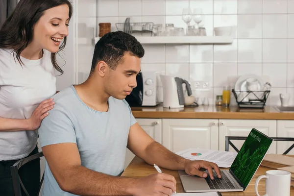 Mujer alegre de pie cerca de novio de raza mixta celebración de pluma cerca de la computadora portátil con el sitio web de comercio en línea - foto de stock