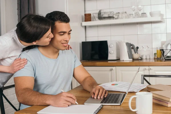 Mulher alegre beijando bochecha de feliz mestiço namorado segurando caneta perto de notebook e laptop, conceito de estudo on-line — Fotografia de Stock