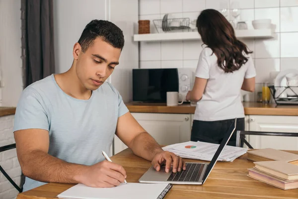 Selective focus of handsome mixed race man writing in notebook near laptop and girl, online study concept — Stock Photo