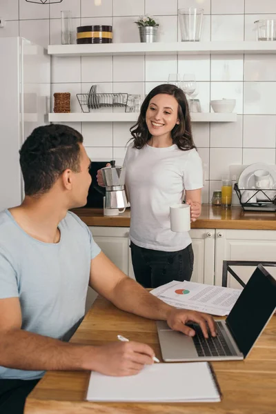 Messa a fuoco selettiva di allegra ragazza in possesso di caffettiera e tazza vicino uomo razza mista e laptop con schermo bianco, concetto di studio online — Foto stock