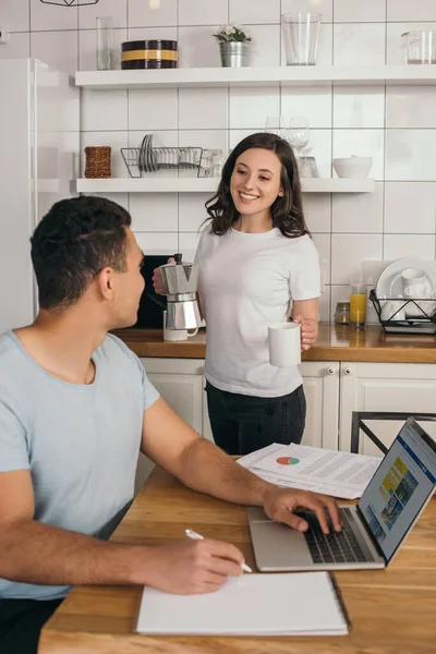 KYIV, UCRANIA - 14 de mayo de 2020: enfoque selectivo de niña alegre sosteniendo cafetera y taza cerca de hombre de raza mixta y portátil con sitio web de reserva - foto de stock