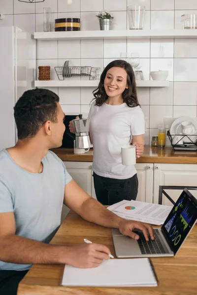 Foco seletivo de menina alegre segurando panela de café e xícara perto de homem de raça mista, laptop com gráficos e gráficos — Fotografia de Stock