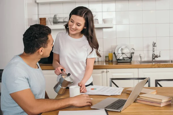 Cheerful girl holding coffee pot and cup near mixed race man and laptop on table — Stock Photo