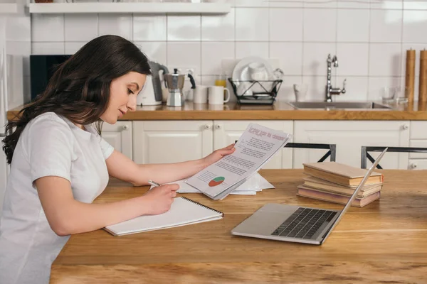 Attractive freelancer holding charts and graphs near laptop while writing in notebook — Stock Photo