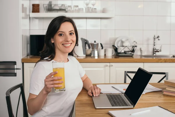 Happy woman holding glass of orange juice near laptop with blank screen — Stock Photo