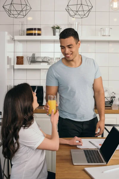 Homem de raça mista feliz dando vidro de suco de laranja para namorada perto de laptop com tela em branco — Fotografia de Stock