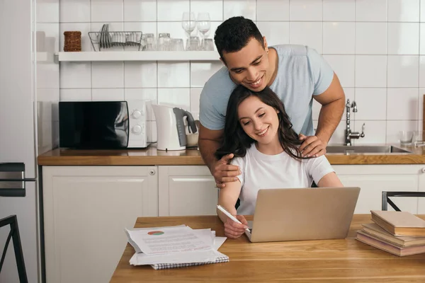 Felice uomo razza mista abbracciando fidanzata allegra e guardando il computer portatile — Foto stock