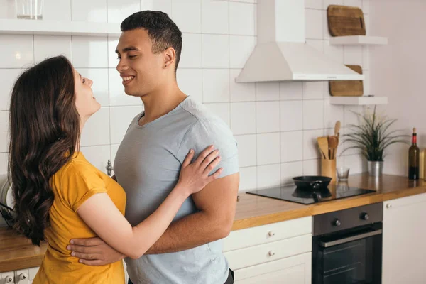 Cheerful mixed race man and smiling girlfriend looking at each other in kitchen — Stock Photo