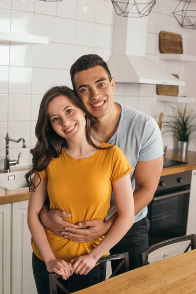 Cheerful mixed race man embracing smiling girlfriend in kitchen — Stock Photo