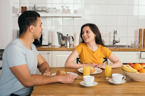 Donna felice guardando fidanzato bi-razziale vicino gustosa colazione sul tavolo — Foto stock