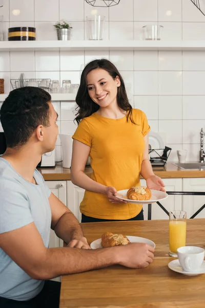 Happy woman holding plate with tasty croissant near mixed race boyfriend — Stock Photo