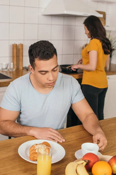 Enfoque selectivo de hombre de raza mixta guapo tocando taza cerca de croissant sabroso, frutas y jugo de naranja en la mesa - foto de stock