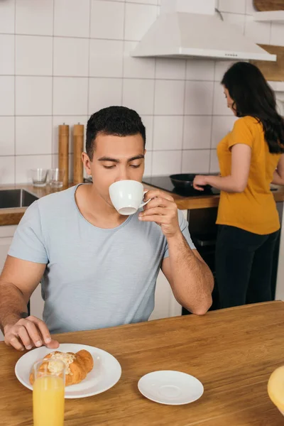 Foco seletivo do homem bi-racial beber café perto da mulher cozinhar em casa — Fotografia de Stock