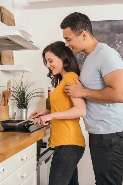 Side view of cheerful mixed race man standing with girlfriend holding wooden spatula near frying pan — Stock Photo