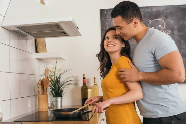 Alegre mixto raza hombre de pie y tocando novia celebración de madera espátula cerca de sartén - foto de stock