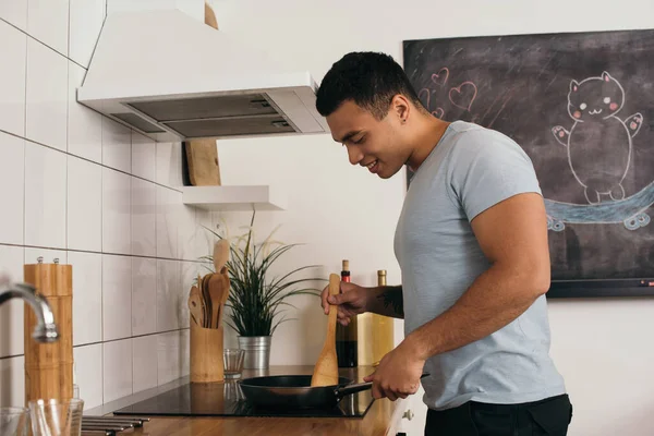 Selective focus of cheerful bi-racial man holding wooden spatula near frying pan — Stock Photo