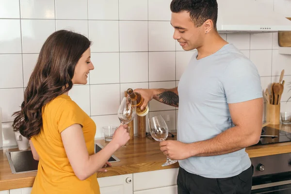 Tattooed mixed race man holding bottle of wine near glasses and cheerful girlfriend — Stock Photo