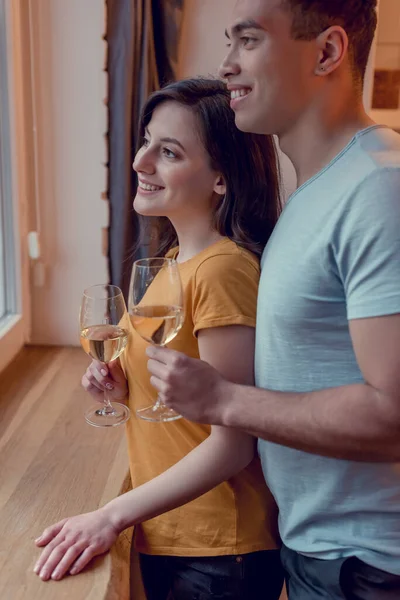 Alegre pareja interracial sosteniendo vasos con vino blanco en casa - foto de stock