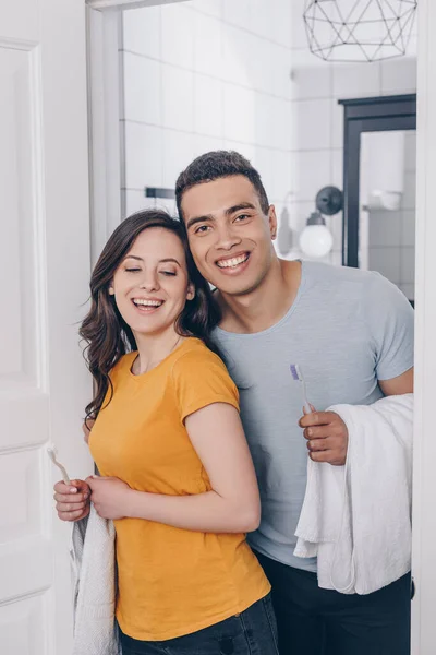 Couple multiracial joyeux tenant serviettes et brosses à dents dans la salle de bain — Photo de stock