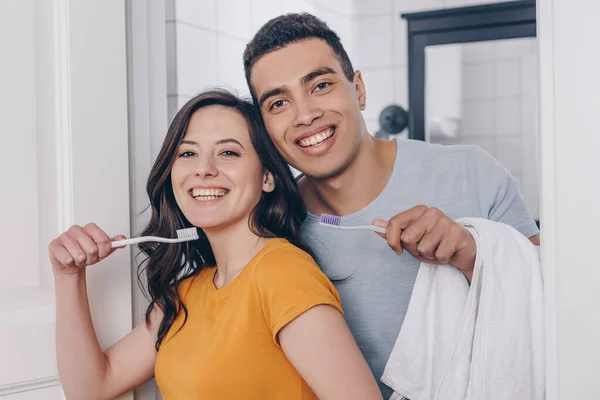 Feliz pareja multirracial sosteniendo cepillos de dientes en el baño - foto de stock