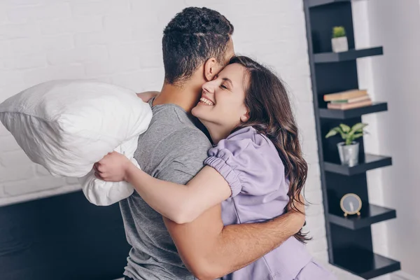 Bela e feliz mulher abraçando misto raça homem enquanto segurando travesseiro — Fotografia de Stock