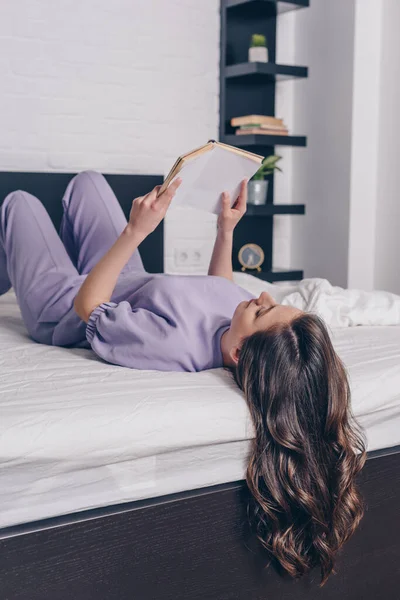 Atractiva joven tumbada en la cama y leyendo un libro - foto de stock