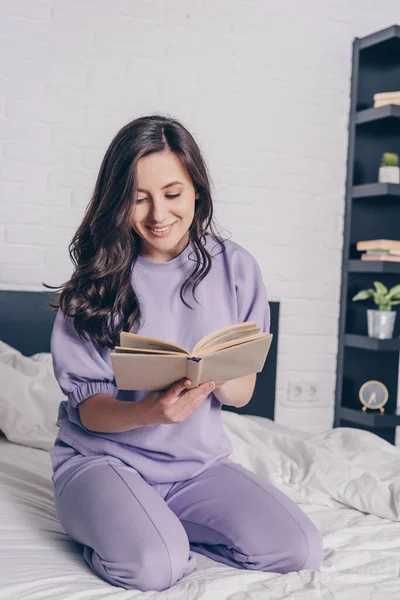 Atractiva joven sonriendo mientras está sentada en la cama y leyendo el libro - foto de stock