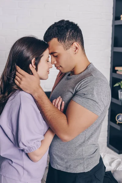 Guapo bi-racial hombre con los ojos cerrados abrazando alegre chica en el dormitorio - foto de stock