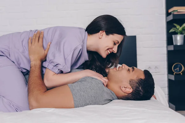 Side view of cheerful woman lying on handsome bi-racial man in bedroom — Stock Photo