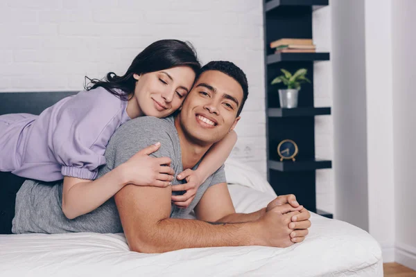 Весела жінка з закритими очима обіймає красивого бі-расового чоловіка, лежачи на ліжку — стокове фото