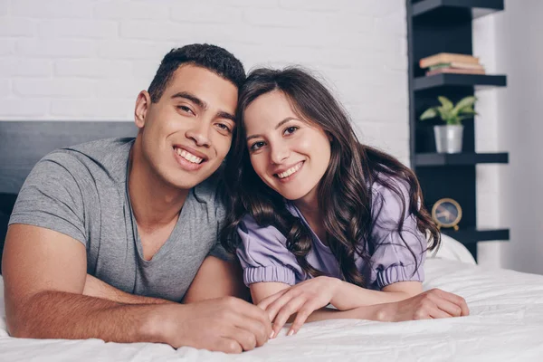 Alegre y joven pareja multirracial sonriendo en el dormitorio - foto de stock