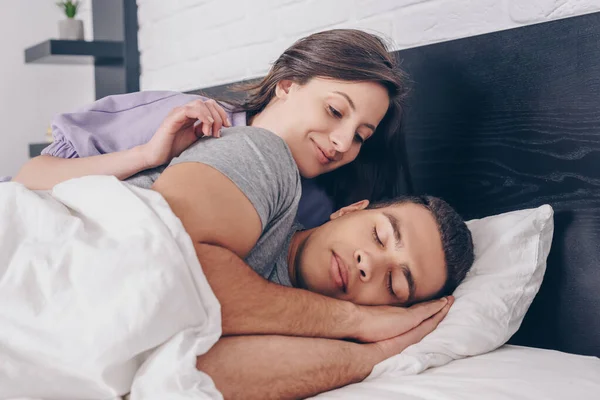 Atractiva mujer mirando guapo mestizo raza hombre durmiendo en dormitorio — Stock Photo