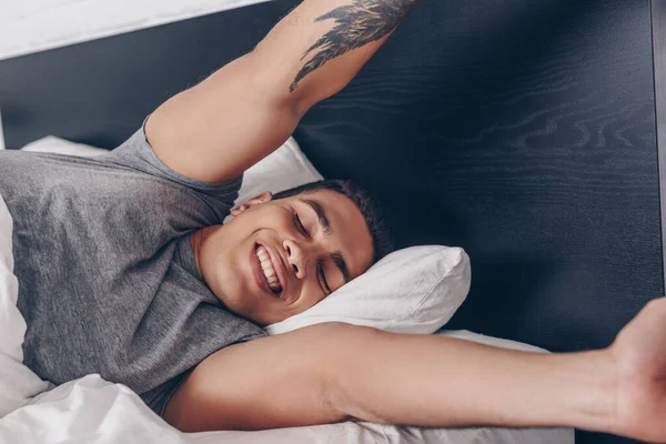 Fröhliche bi-rassische Mann mit Tätowierung Stretching im Bett — Stockfoto
